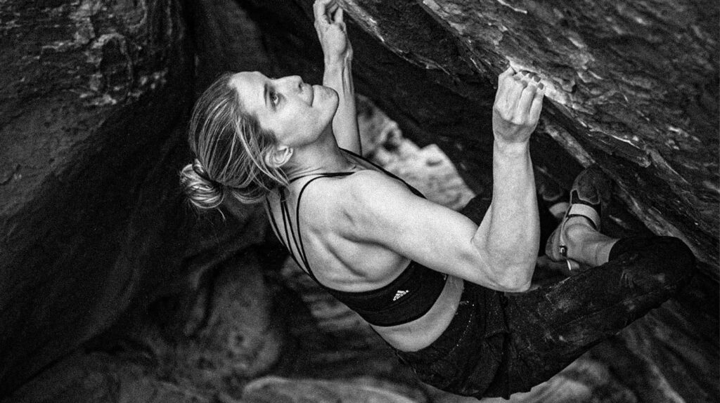 A black and white photo of a woman rock climbing, gripping an overhanging boulder focused and determined.