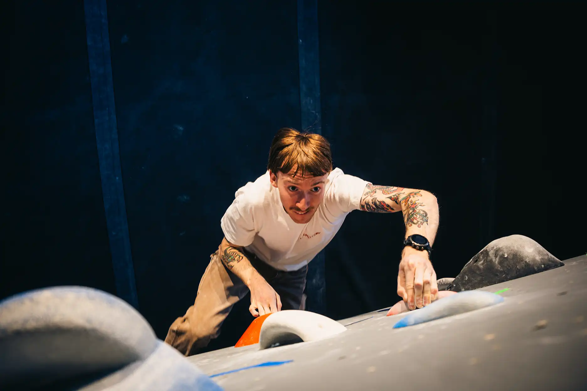 A person wearing a white shirt is climbing an indoor rock wall, gripping holds with both hands and looking upward.