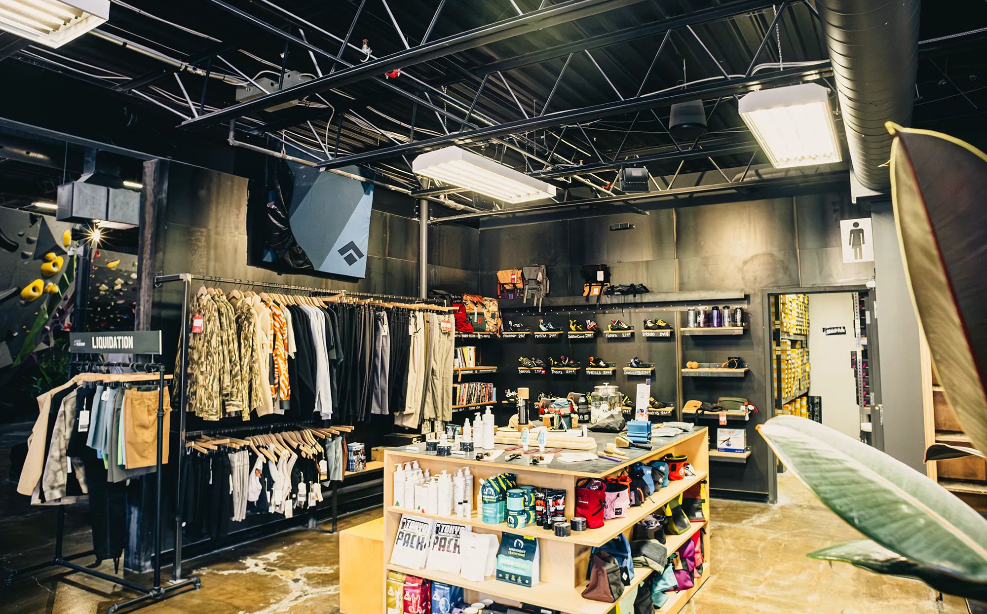 Clothing and gear store interior with racks of clothes, shelves of accessories, and an island display of products. Industrial design with exposed ceiling and bright lighting.