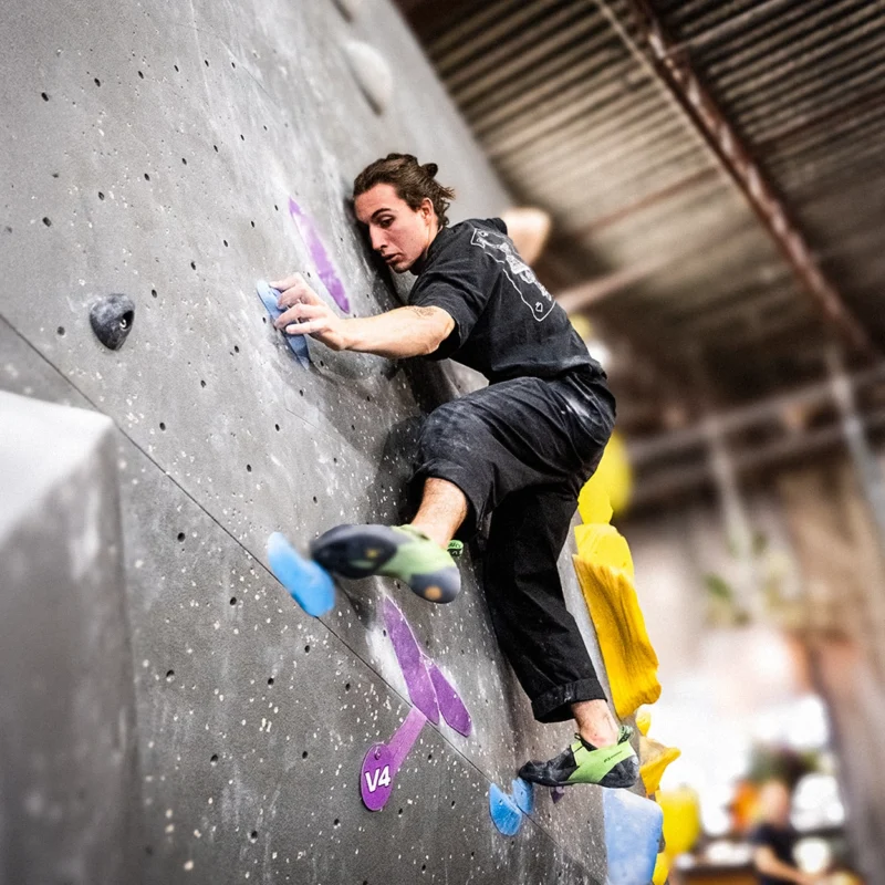 A person is climbing an indoor bouldering wall, gripping holds with hands and feet. The wall features various colored holds and is marked with a "V4" level tag.