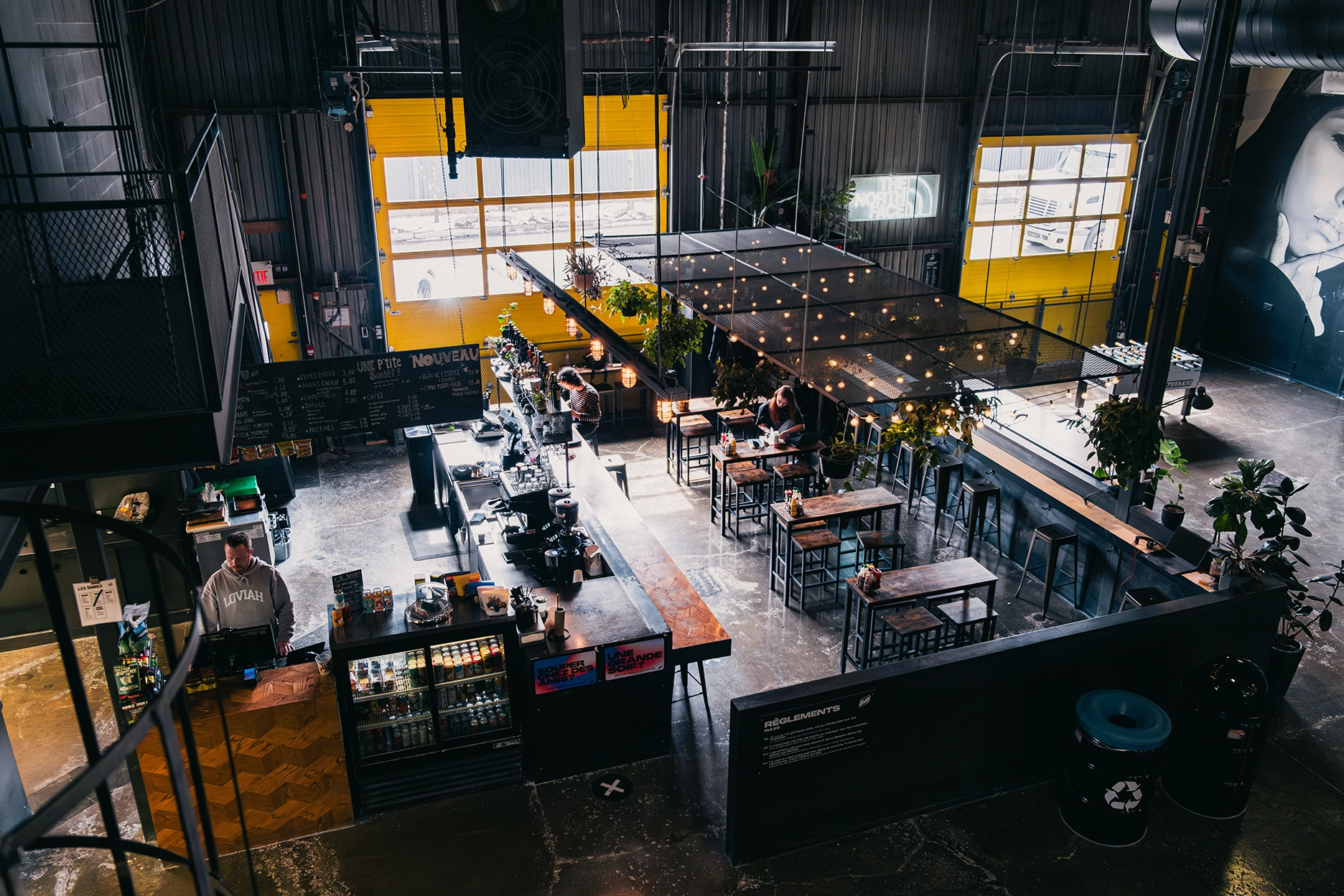 A modern industrial-style café with large windows, string lights, high ceilings, multiple tables, and people sitting and ordering at the counter.