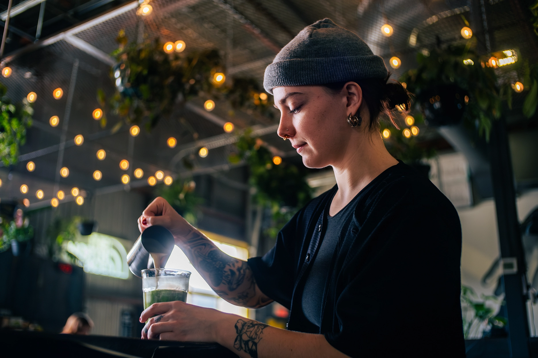 Une personne portant un bonnet gris verse du café d'un petit pichet dans un verre dans un bar ou un café avec des lumières suspendues et des plantes.