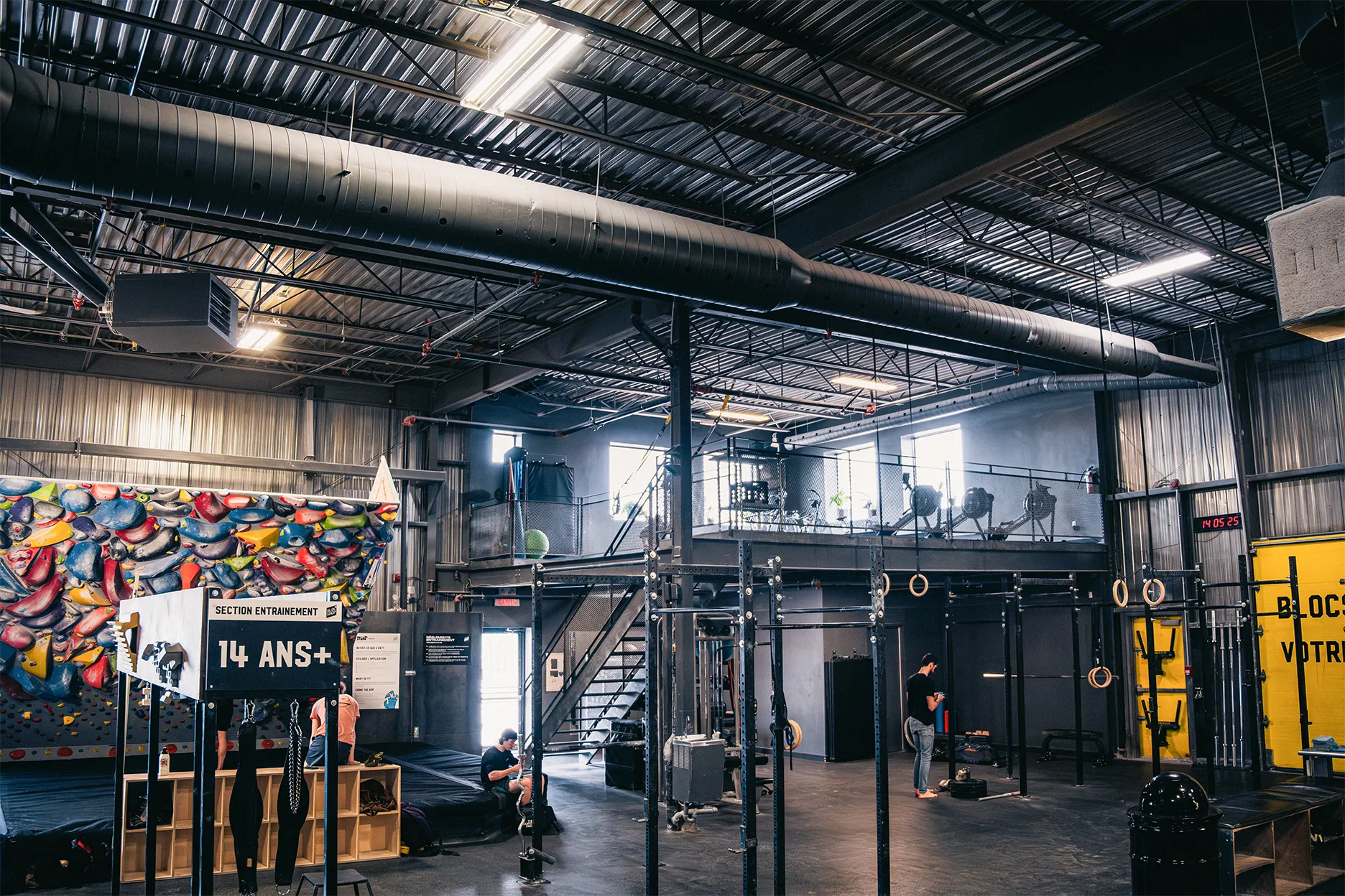 Industrial-style gym interior with exposed metal beams, climbing wall, gym equipment, rings, and a staircase leading to a mezzanine level. Several people are exercising.