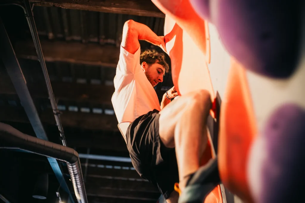 A person wearing a white shirt and black shorts climbs an indoor rock climbing wall with purple holds.