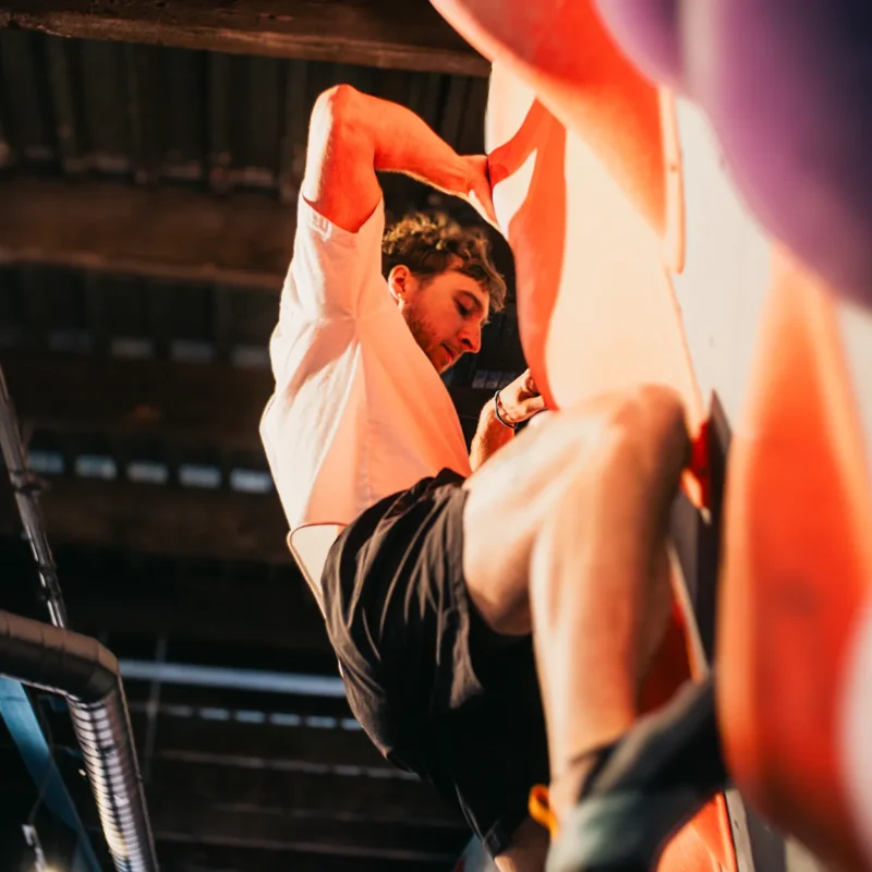 A person wearing a white shirt and black shorts climbs an indoor rock climbing wall with purple holds.