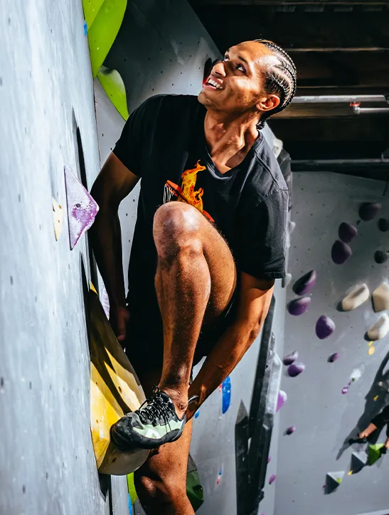 A person in athletic gear climbs an indoor bouldering wall at one of the best bouldering centers in Montreal, gripping holds with one hand and foot, while looking upward and smiling.