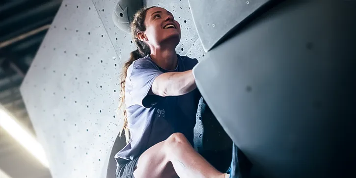 At one of the best bouldering centers in Montreal, a woman climbs an indoor rock wall, gripping handholds and looking upward with a focused expression.