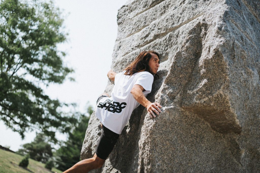 A person with long hair climbs a large rock outdoors, wearing a white T-shirt and black shorts. Trees and grass are visible in the background.