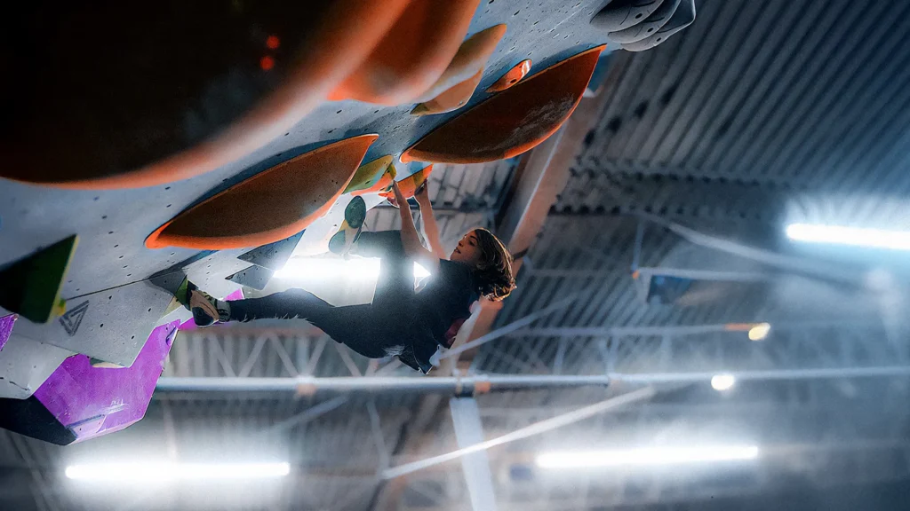 A person climbs an indoor bouldering wall with large orange holds, suspended upside down under artificial lighting.
