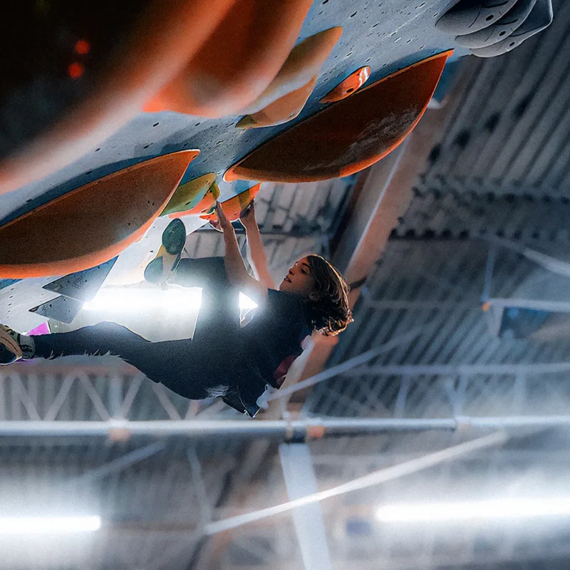 A person climbs an indoor bouldering wall with large orange holds, suspended upside down under artificial lighting.
