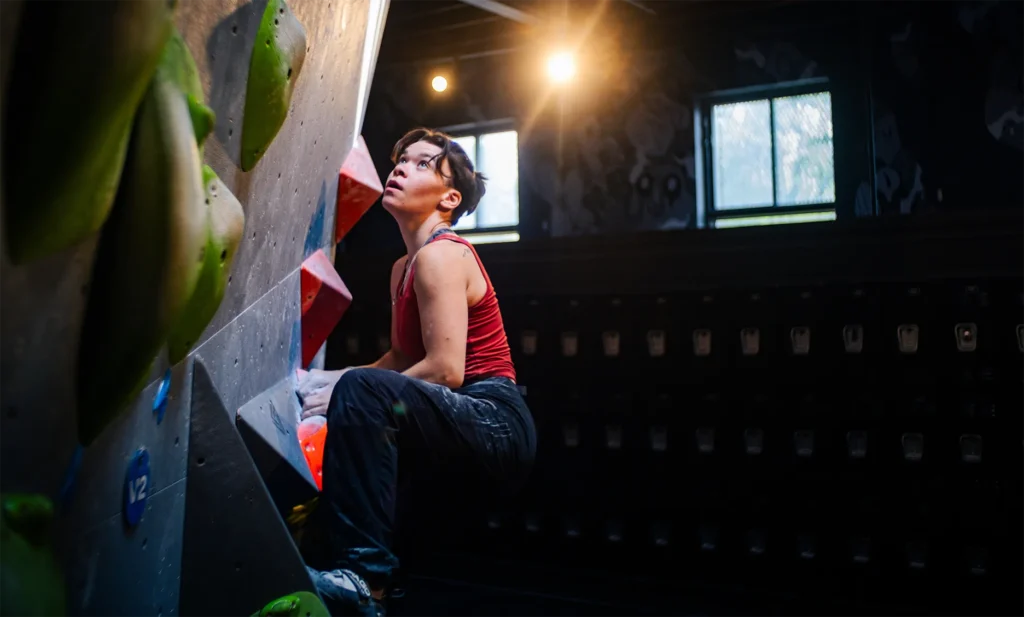 A person in a red tank top climbs an indoor bouldering wall, looking up toward the next handhold under bright lights.