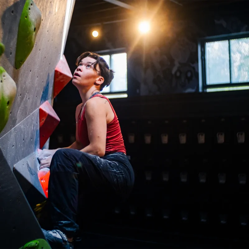 A person in a red tank top climbs an indoor bouldering wall, looking up toward the next handhold under bright lights.