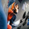 A woman climbs an indoor bouldering wall, reaching for a hold with her left hand and balancing on large orange holds.