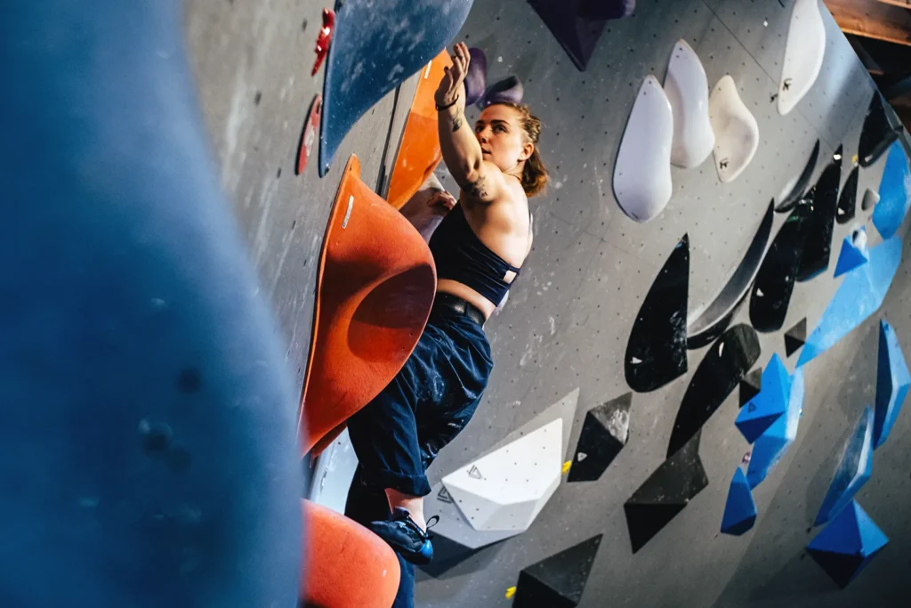 Une personne en tenue de sport escalade un mur d'escalade en salle avec des prises colorées de différentes formes et tailles.
