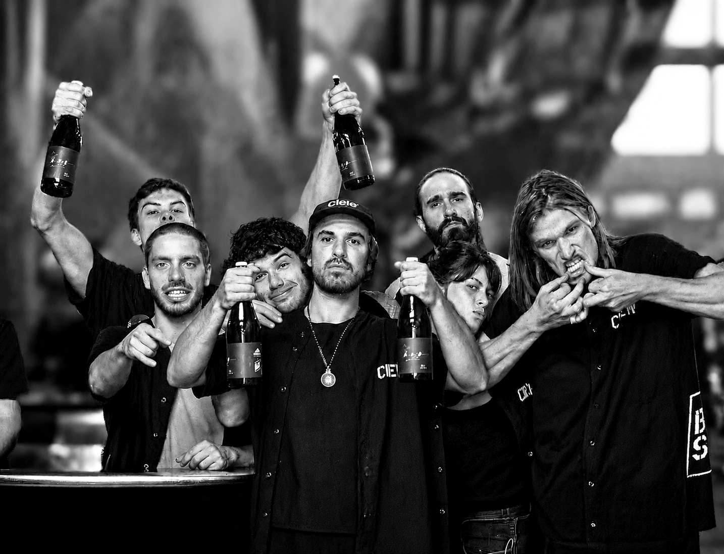 Un groupe de sept personnes pose ensemble, tenant des bouteilles et faisant des gestes expressifs, dans un décor en noir et blanc qui semble être un bar ou un espace industriel.