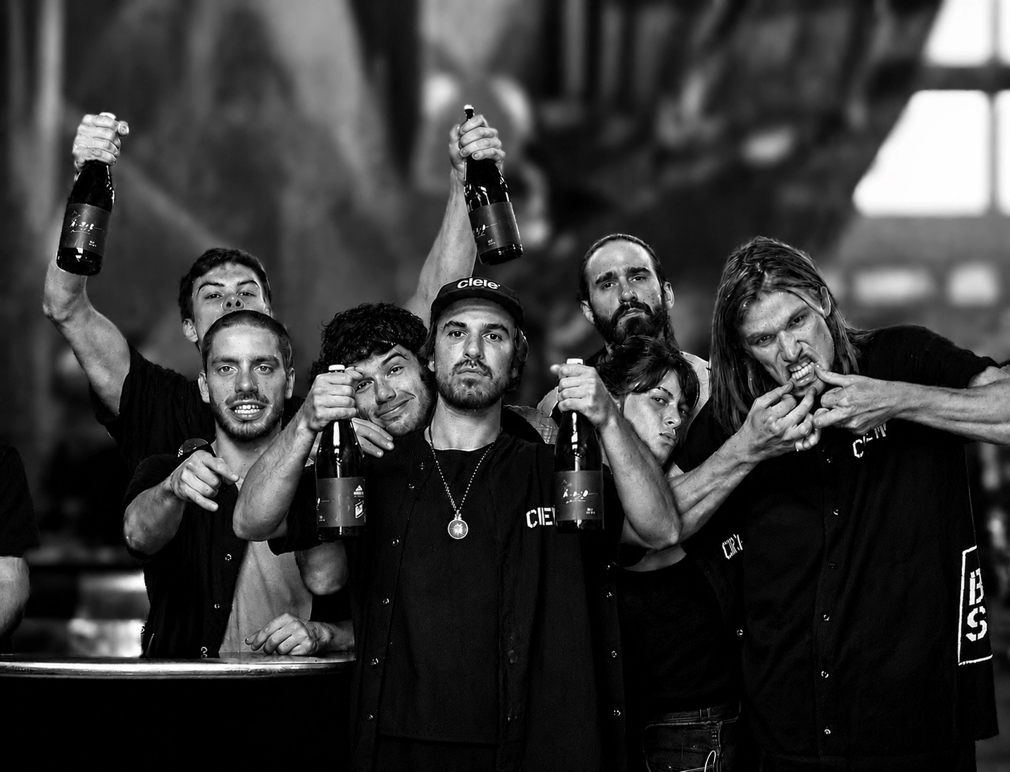 A group of seven people in matching shirts hold and display champagne bottles, with some pointing or posing for the camera in a celebratory manner.